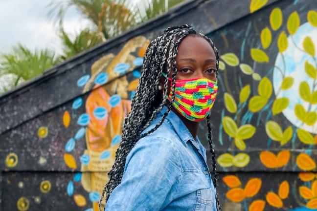 A woman wearing a colorful knit face mask turns to face the camera