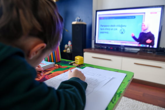 A young girl works on a school assignment with a teacher visible on her television