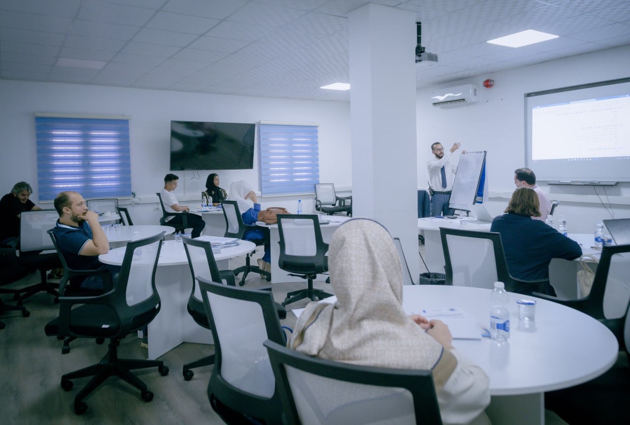 La photo montre 8 personnes, hommes et femmes, dans une salle de réunion face à un homme qui fait une présentation à l’aide d’un écran.