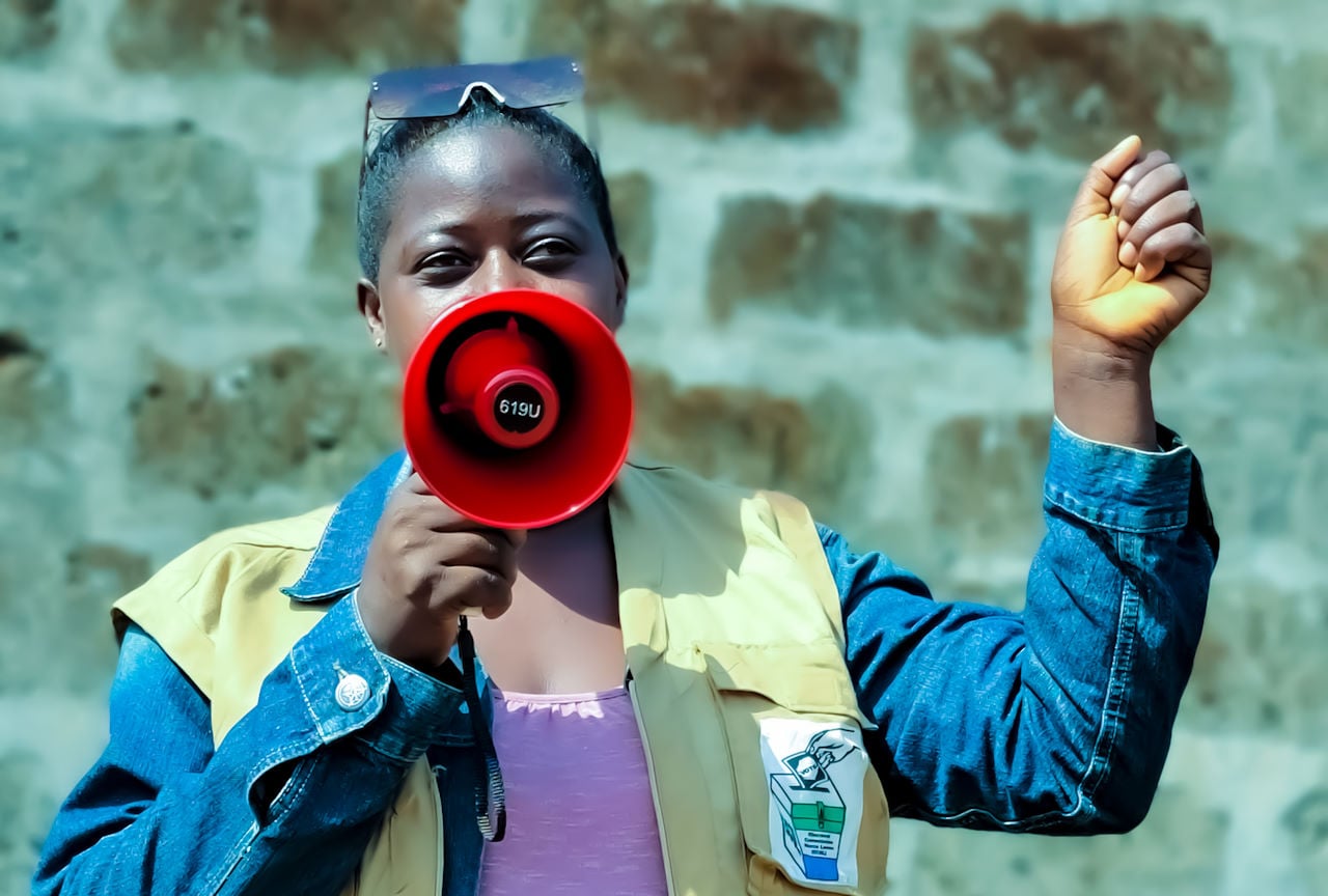Une femme en extérieur tient un mégaphone et fait un discours, le point levé.